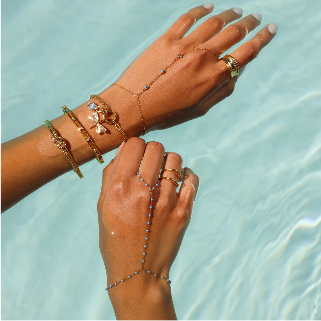 Close up image of a model's wrist showcasing a gold bracelet stack and beaded hand chains over a water background