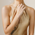 Woman wearing a beige halter top with tattoos on her arms, holding her hand to her neck and showcasing a dainty floral diamond bracelet.