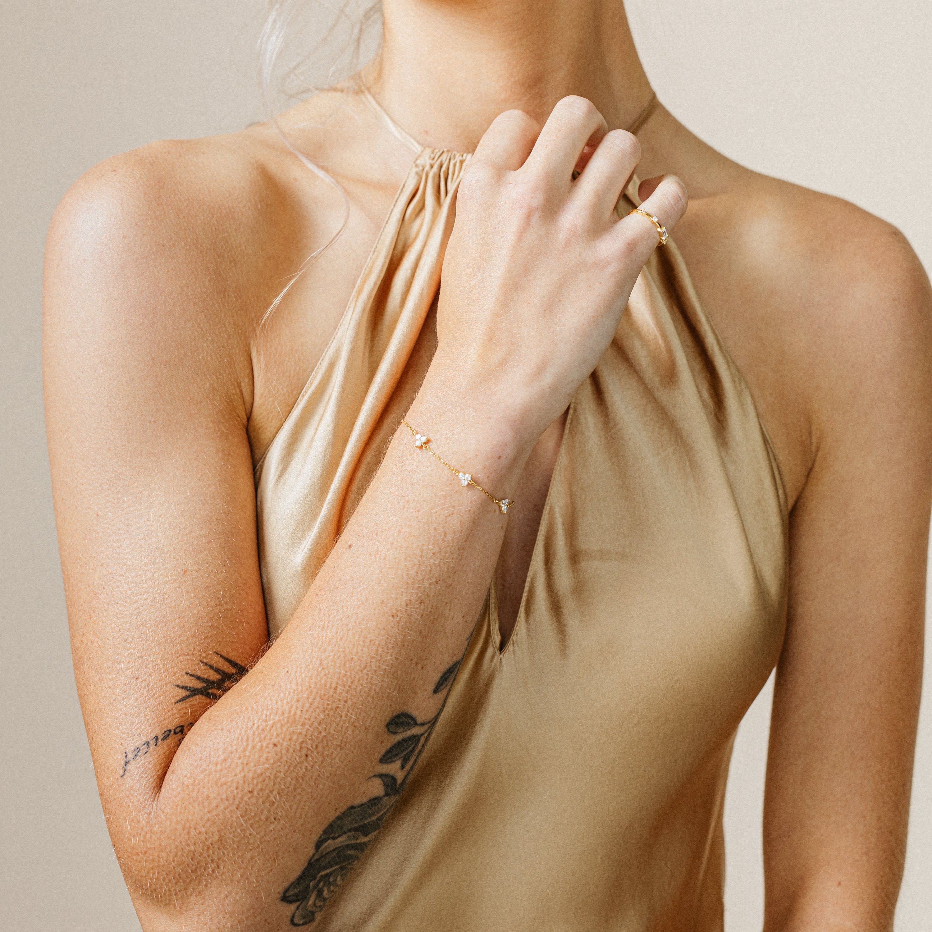 Woman wearing a beige halter top with tattoos on her arms, holding her hand to her neck and showcasing a dainty floral diamond bracelet.