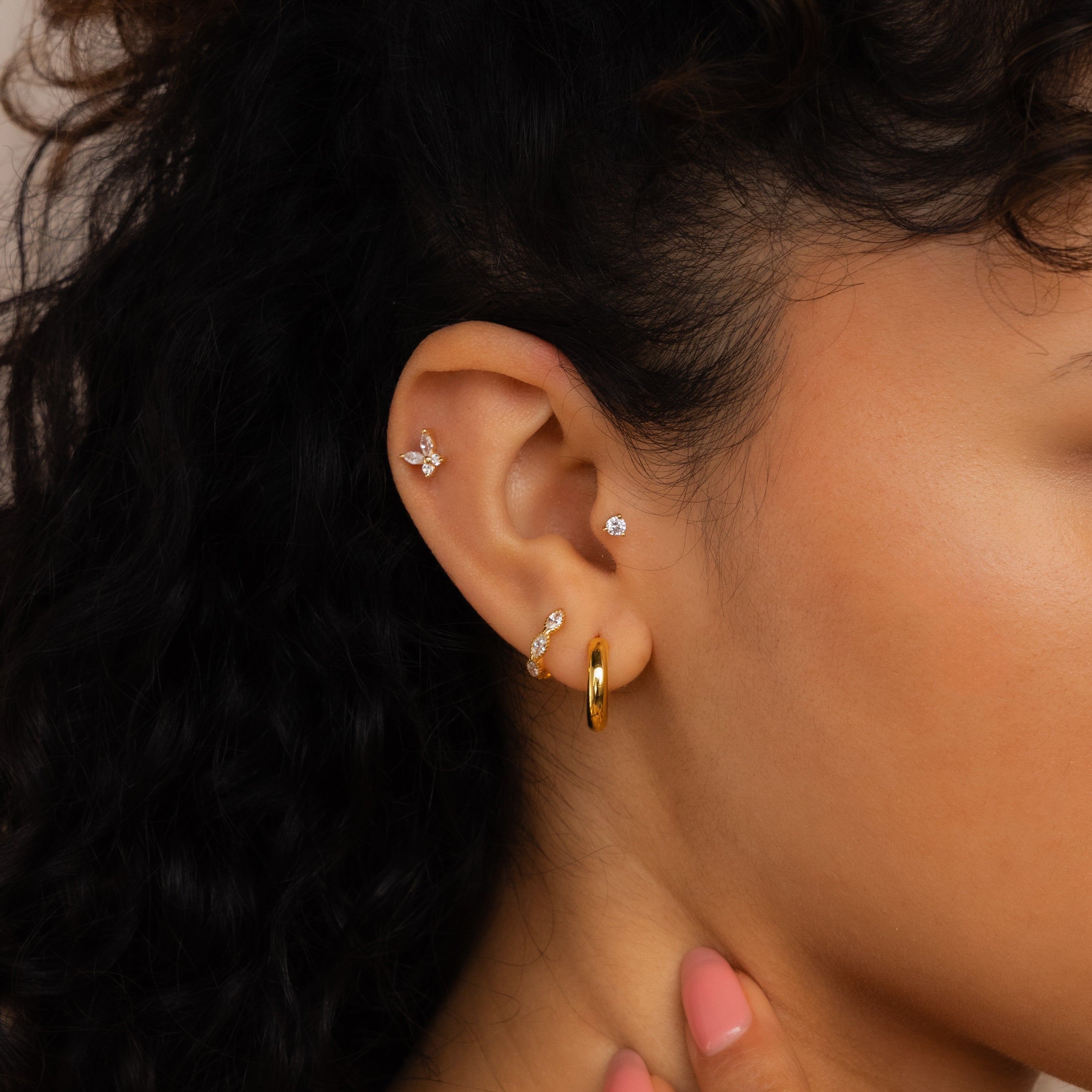 Close-up of a woman's ear adorned with the Diamond Butterfly Earrings Set and other gold and diamond pieces. Her hand rests by her chin, highlighting a Layering Jewelry style, with curly hair visible in the shot.