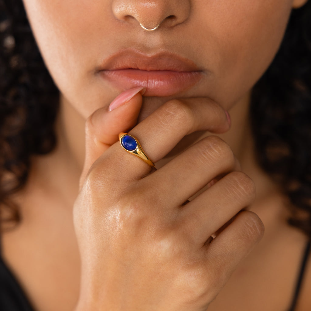A woman with curly hair and a nose ring wears the Signet Mood Ring, featuring a blue color-changing stone, her hand thoughtfully near her lips.