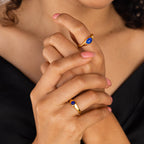 A woman with pink nails wears two Signet Mood Rings, featuring color-changing stones and blue gems, on her fingers against a black top.
