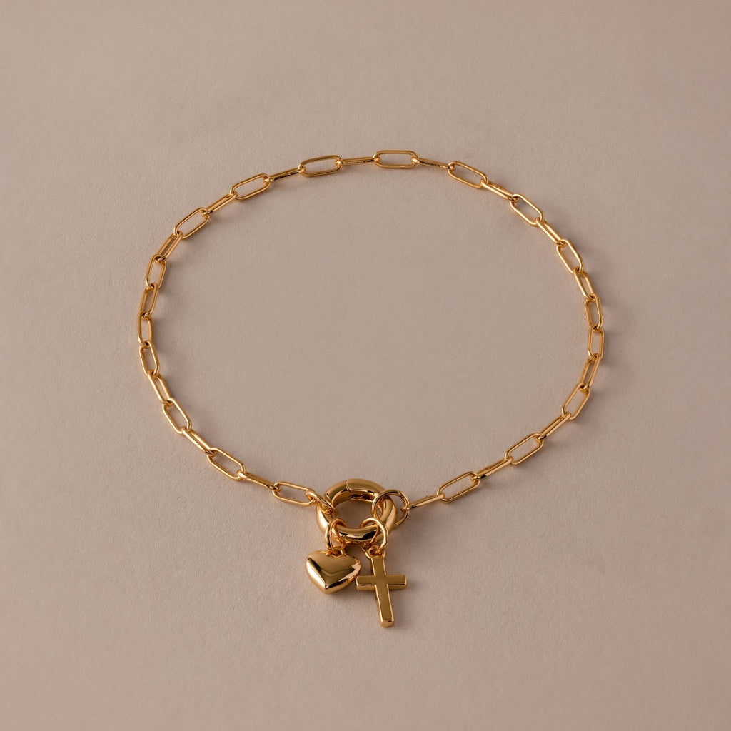 The Cross & Heart Charm Bracelet features a minimalist gold paperclip chain with delicate cross and heart pendants, beautifully displayed on a beige background.