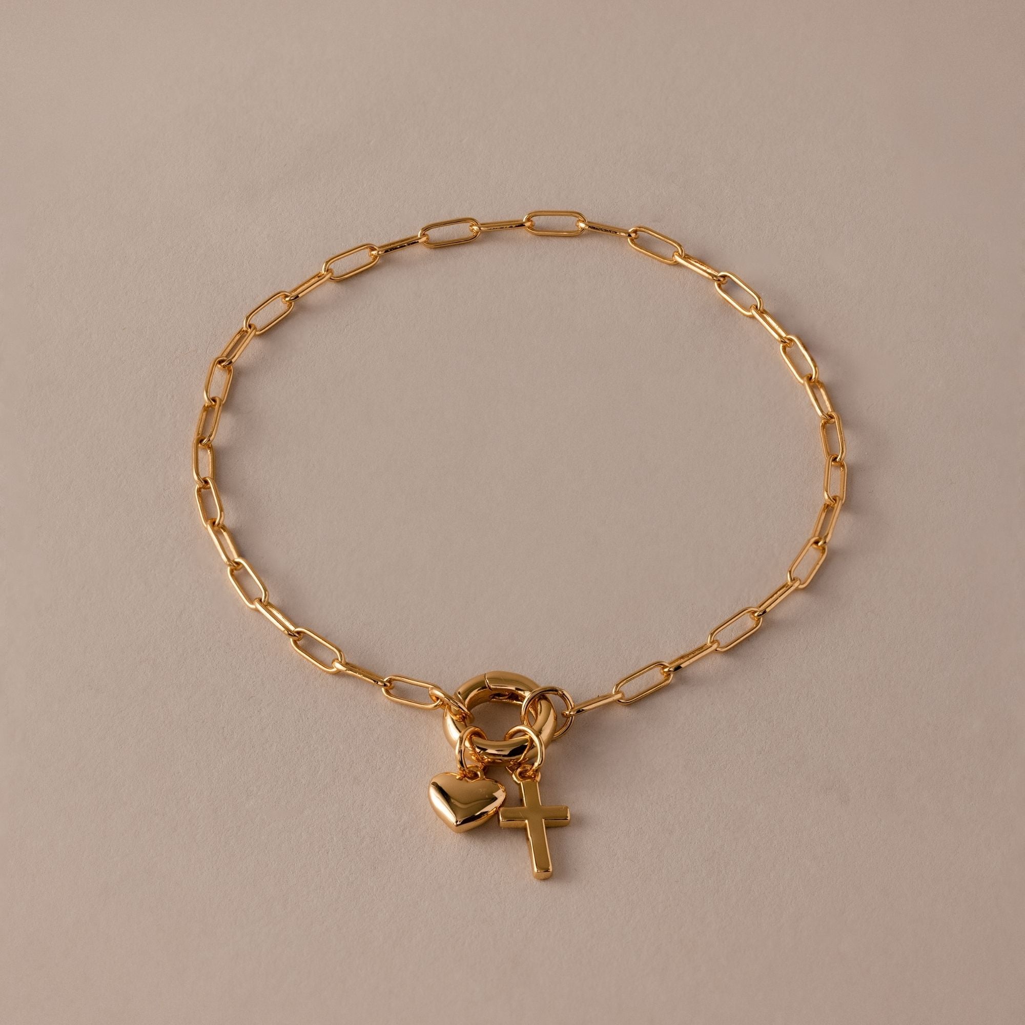 The Cross & Heart Charm Bracelet features a minimalist gold paperclip chain with delicate cross and heart pendants, beautifully displayed on a beige background.