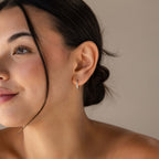 A close-up of a woman's ear wearing our Classic Diamond Sprinkle Huggie earring with a small vertical clear stone accent, set against a soft beige background.