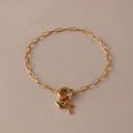 The Cross & Heart Charm Bracelet in 18K Gold features a minimalist cross and heart on a gold paperclip chain, displayed on a beige background.