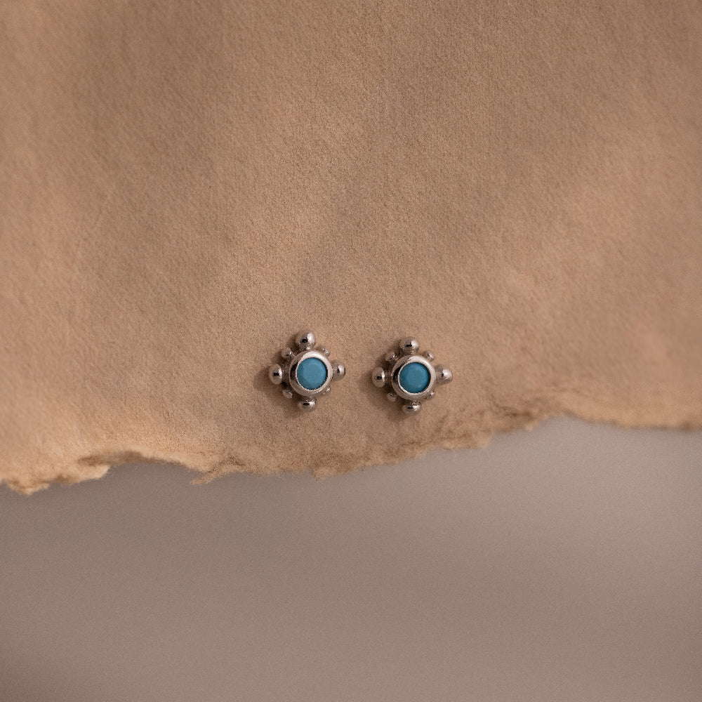 Pair of small turquoise gemstone stud earrings with a delicate silver beaded setting displayed on textured neutral fabric, showcasing a vintage inspired minimalist design.