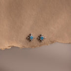 Pair of small turquoise gemstone stud earrings with a delicate silver beaded setting displayed on textured neutral fabric, showcasing a vintage inspired minimalist design.