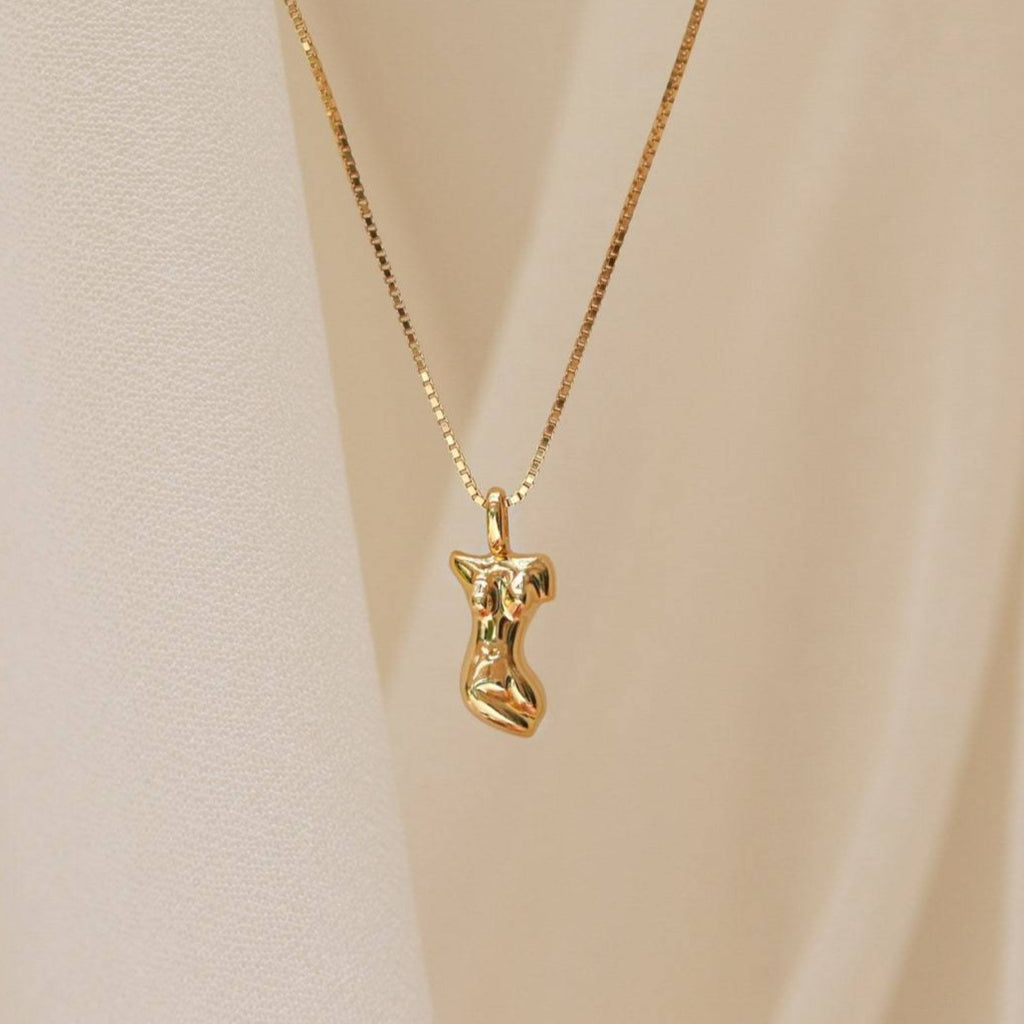 The Kinn Body Necklace features a pendant shaped like a stylized female torso, shown against soft beige fabric for a bold, statement jewelry look.