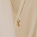 The Kinn Body Necklace features a pendant shaped like a stylized female torso, shown against soft beige fabric for a bold, statement jewelry look.