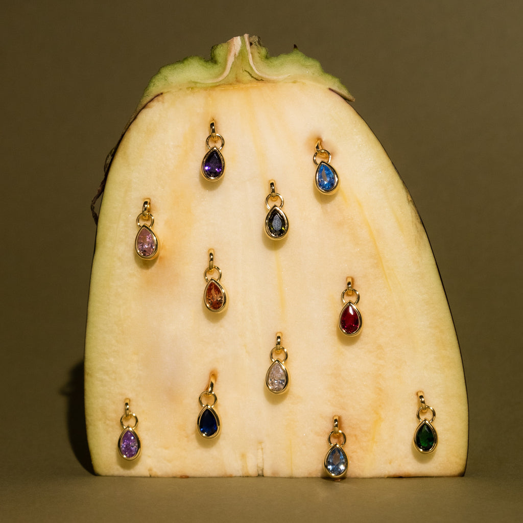 A sliced banana stands upright with twelve Teardrop Birthstone Charms, each in a gold bezel setting, pinned on its flat surface.