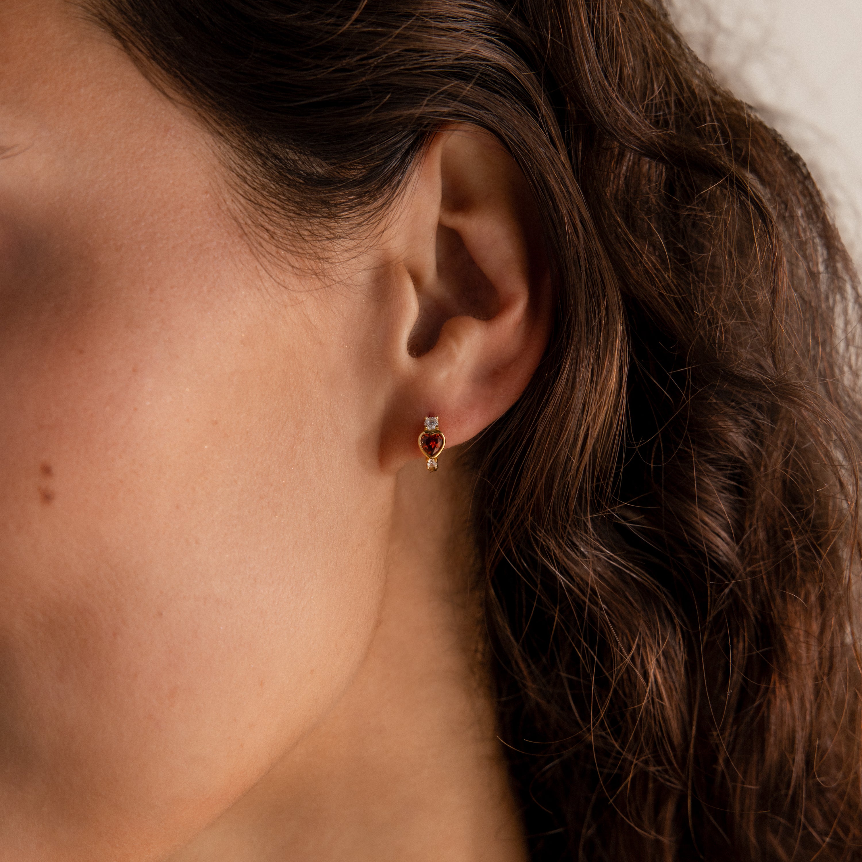 Close-up of a woman's ear with wavy brown hair, wearing Garnet Heart Hoops—small red heart-shaped earrings with two round diamonds on each side