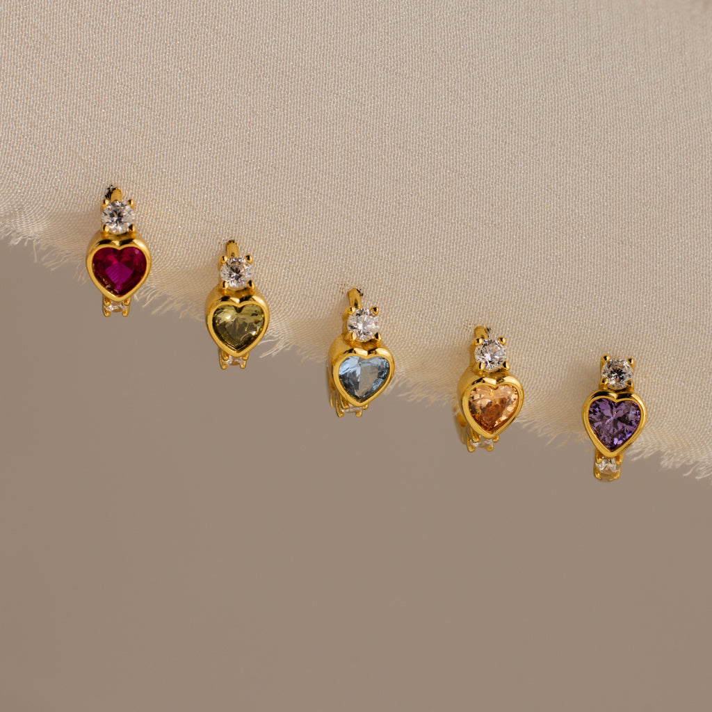 The Birthstone Heart Hoops by Caitlyn Minimalist feature heart-shaped gemstones in various colors, each accented with a clear stone—ideal for pairing with your favorite birthstone earrings or diamond huggies for a coordinated look.
