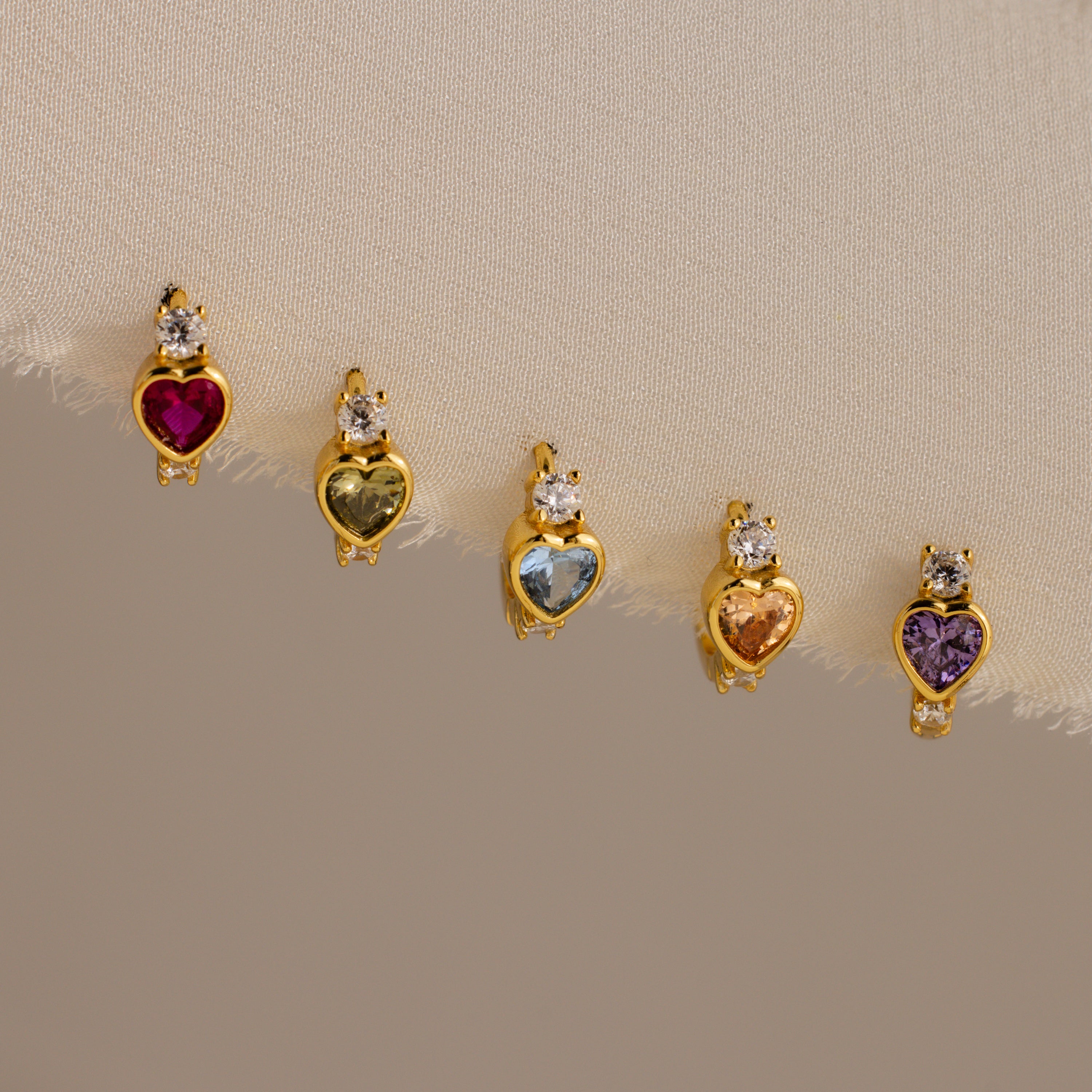 The Birthstone Heart Hoops by Caitlyn Minimalist feature heart-shaped gemstones in various colors, each accented with a clear stone—ideal for pairing with your favorite birthstone earrings or diamond huggies for a coordinated look.