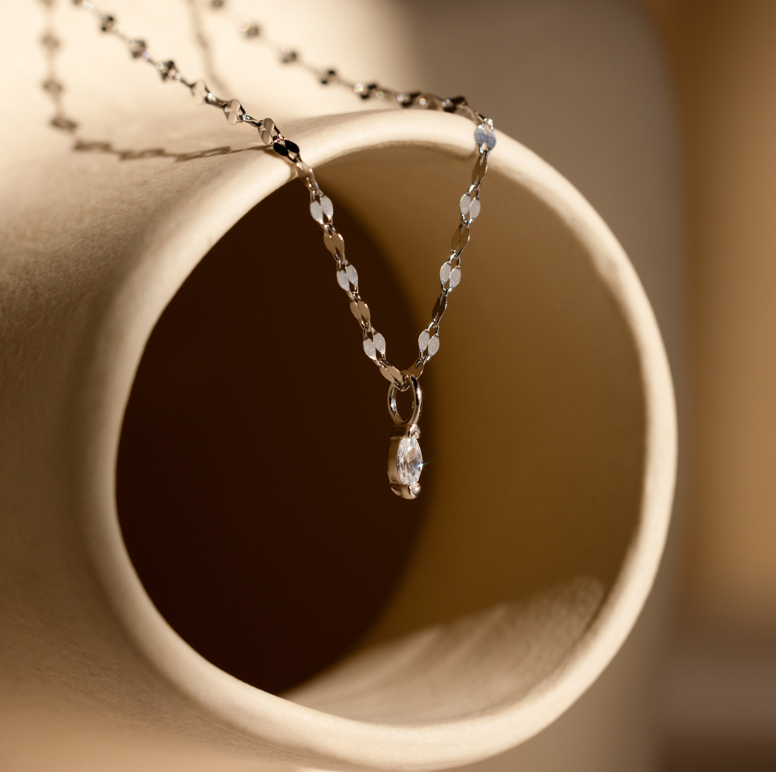 The Dainty Marquise Pendant Necklace in Silver features a slim mirror twist chain and elegant marquise gemstone pendant, artfully displayed atop a beige ceramic tube.