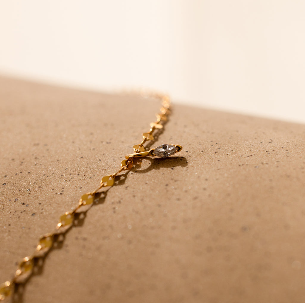 A Dainty Marquise Pendant Necklace in 18K Gold is displayed on a textured beige surface.