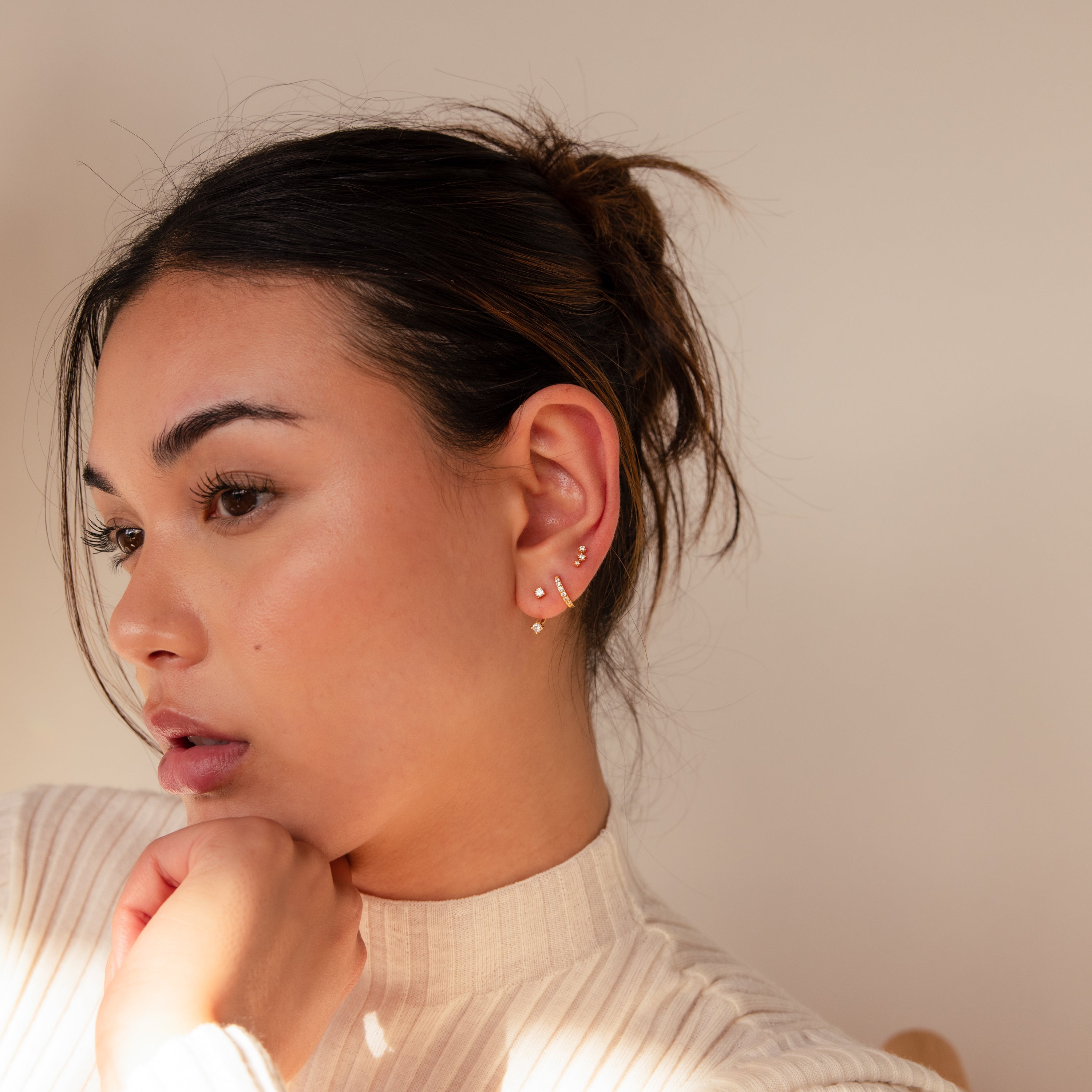 A woman wearing multiple gold earrings, including the Diamond Earrings Set in 18K Gold, rests her chin on her hand and gazes thoughtfully to the side.