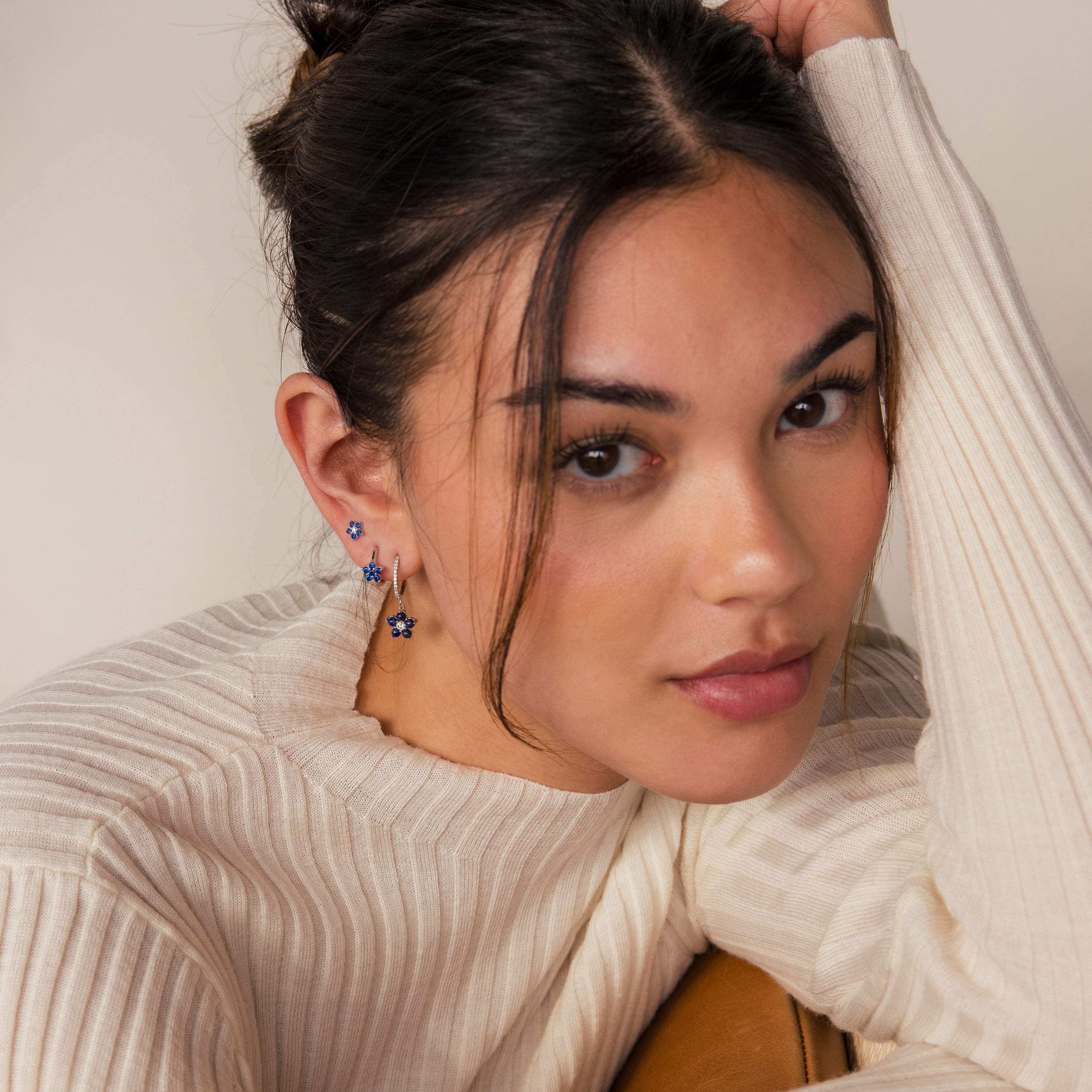 A woman with natural makeup, dressed in a cream blouse, rests her chin on her hand while wearing the Sapphire Flower Earrings Set in silver.