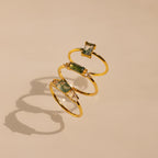 Three gold rings, including the Agate Statement Ring in 18K Gold and a CZ diamond-adorned ring, are artistically displayed in a floating spiral on a beige background.