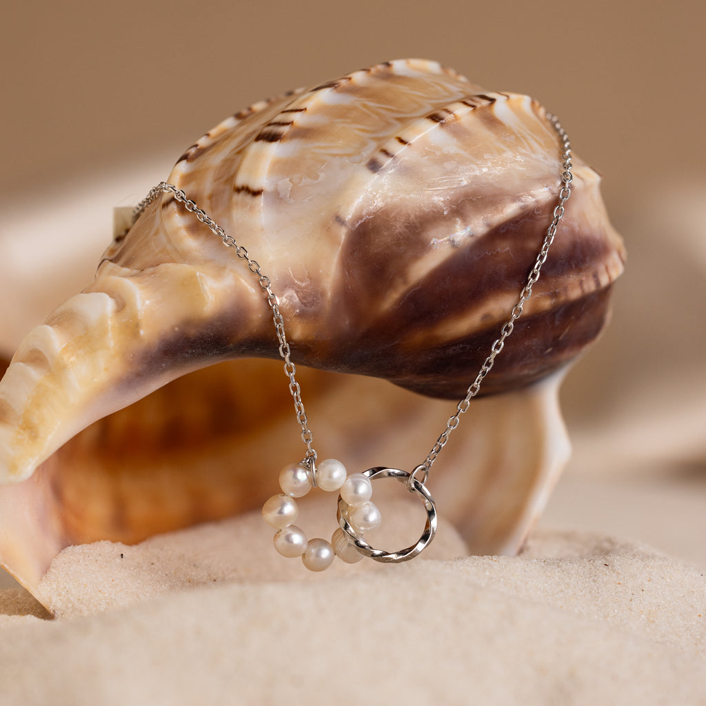 The Interlocking Pearl Necklace in Silver is displayed draped over a seashell on a sandy beach.