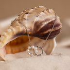 The Interlocking Pearl Necklace in Silver is displayed draped over a seashell on a sandy beach.