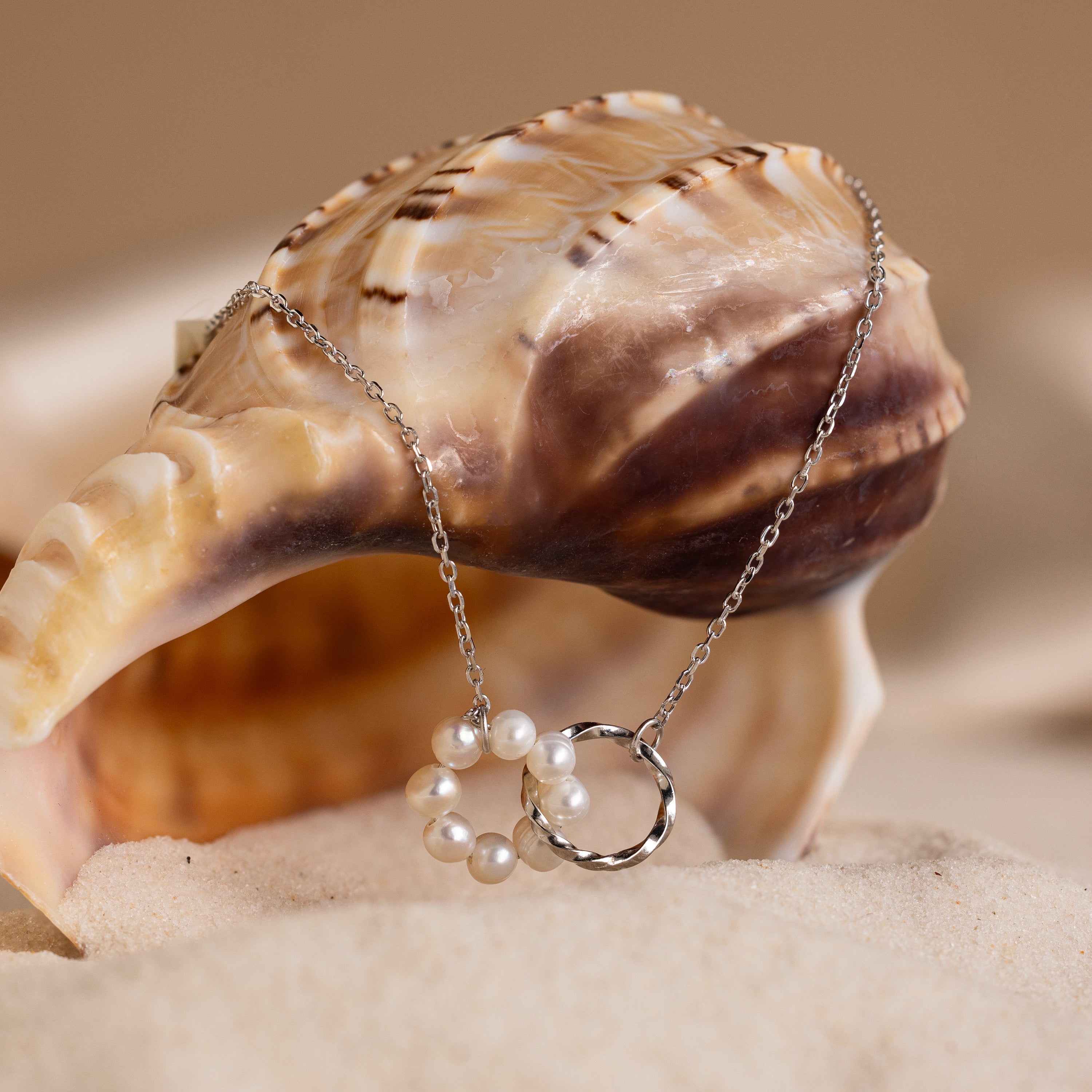 The Interlocking Pearl Necklace in Silver is displayed draped over a seashell on a sandy beach.