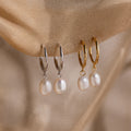 Two pairs of gold and silver hoop earrings with pearl drops on a beige fabric background