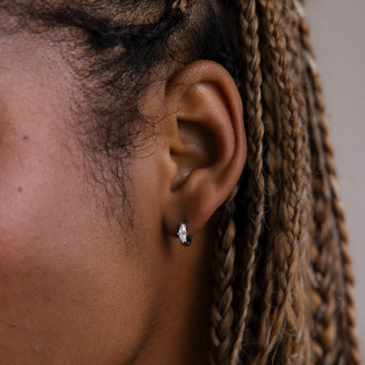 Close-up of an ear with braided hair wearing a small silver hoop earring against a neutral background