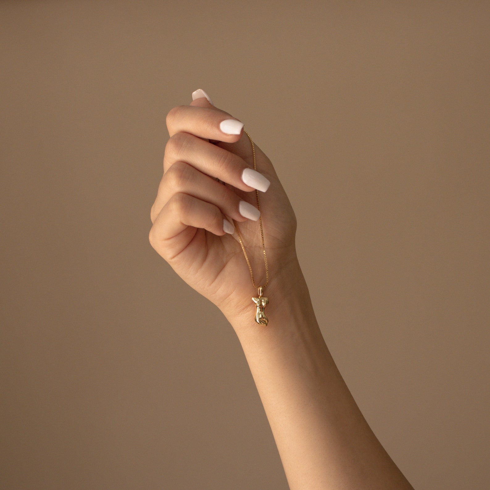 A manicured hand holds the Kinn Body Necklace—a gold body pendant—showcasing this elegant statement jewelry against a beige background.