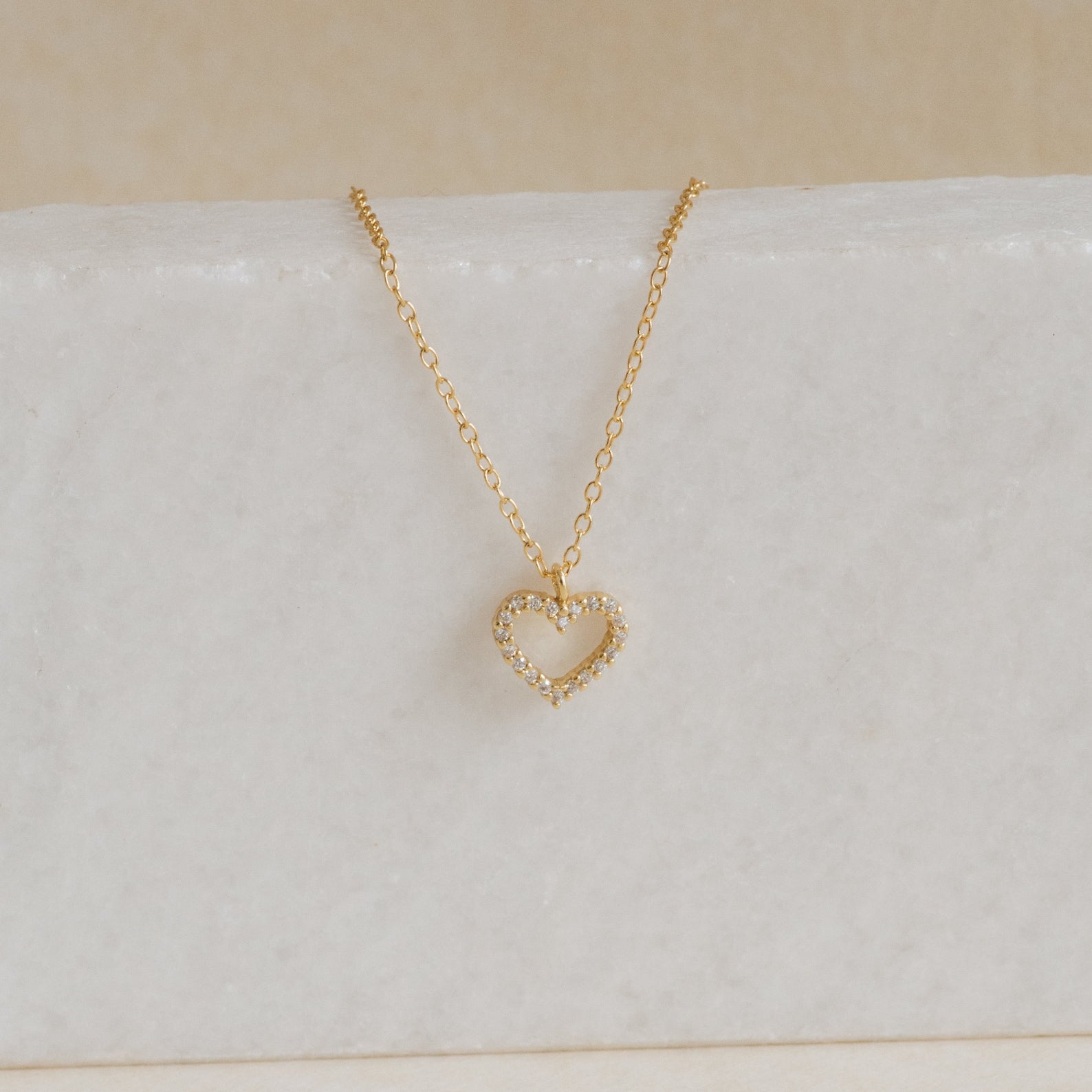 The Forever Pave Heart Necklace features a small diamond-encrusted heart pendant on a gold chain, ideal for elegant bridal jewelry, shown on white marble.
