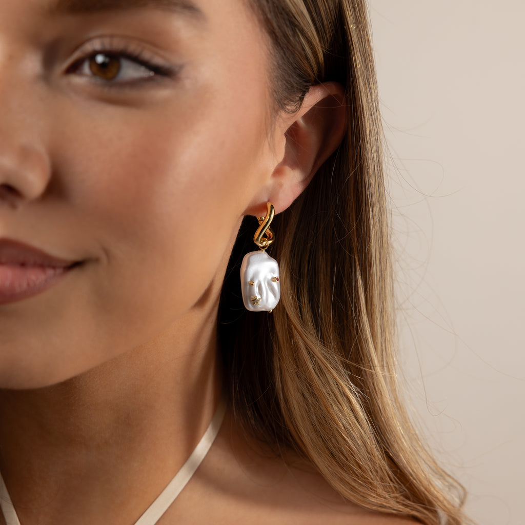 A woman with long brown hair and natural makeup wears Celestial Baroque Pearl Charm Hoops.