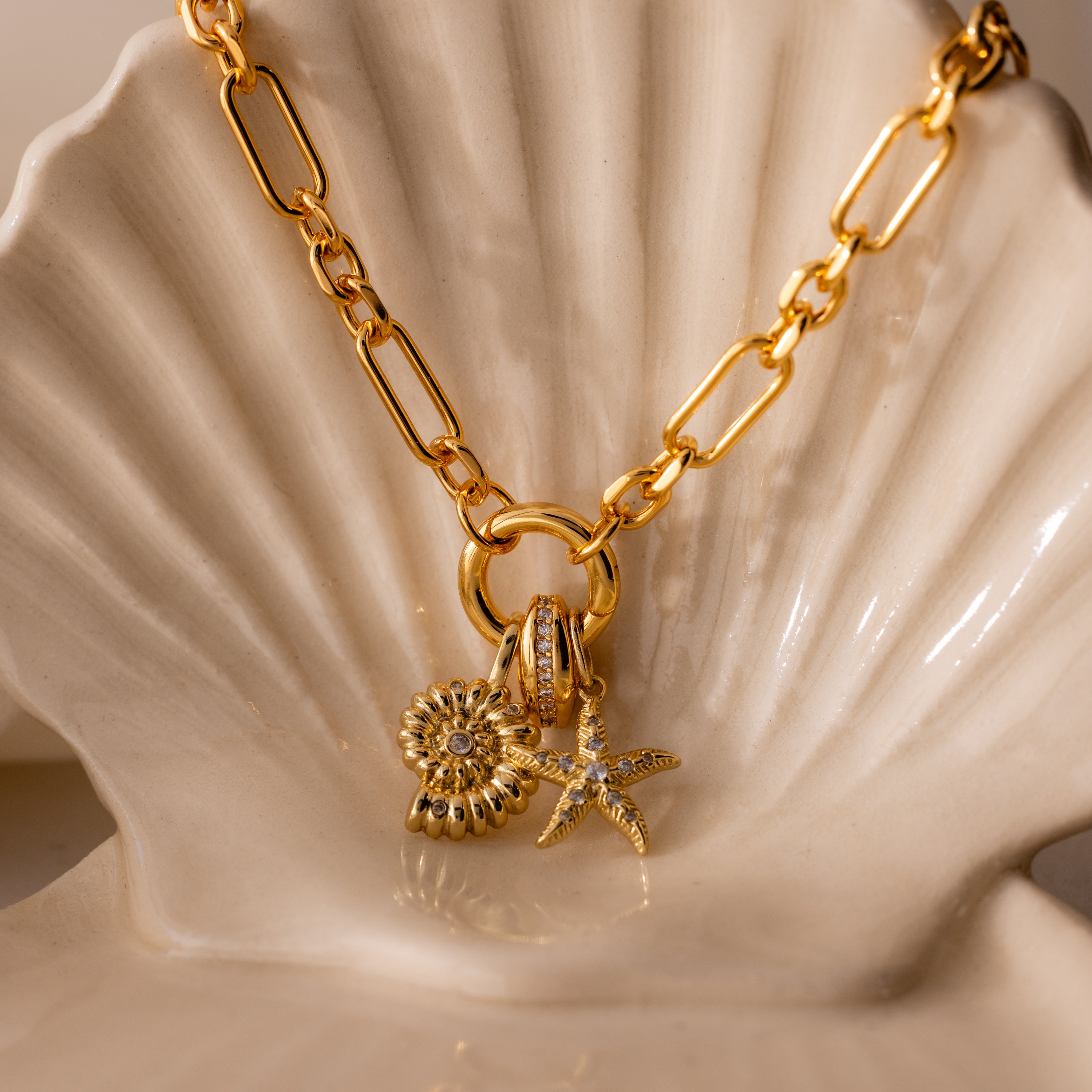 The Pave Dome Charm in 18K Gold adds sparkle, shown stacked with flower and starfish charms on a gold chain necklace, displayed on a white ceramic shell dish.