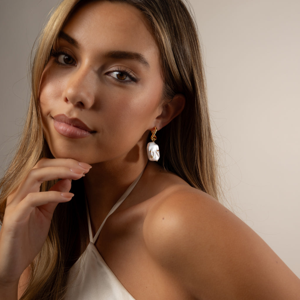 Woman with long hair wearing a white halter top and Celestial Baroque Pearl Charm Hoops in 18K Gold, posing with her hand near her chin.