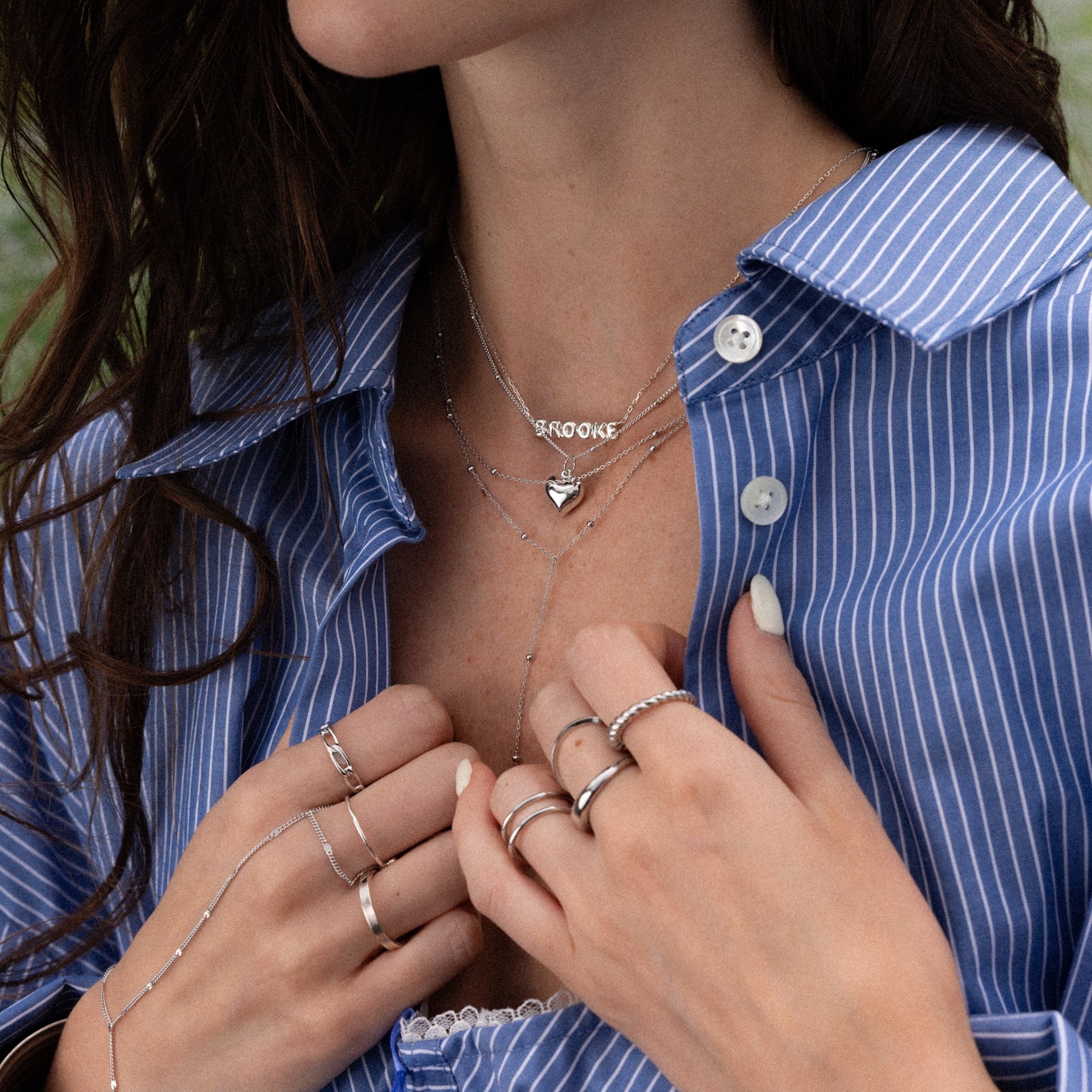 Woman in blue pinstripe shirt wears minimal jewelry: delicate chain rings, layered silver necklaces, and a Duo Satellite Lariat Necklace in Silver.