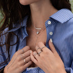 A woman in a blue pinstripe shirt wears the Duo Satellite Lariat Necklace layered with minimal necklaces and several silver rings, some featuring sparkling CZ diamonds.