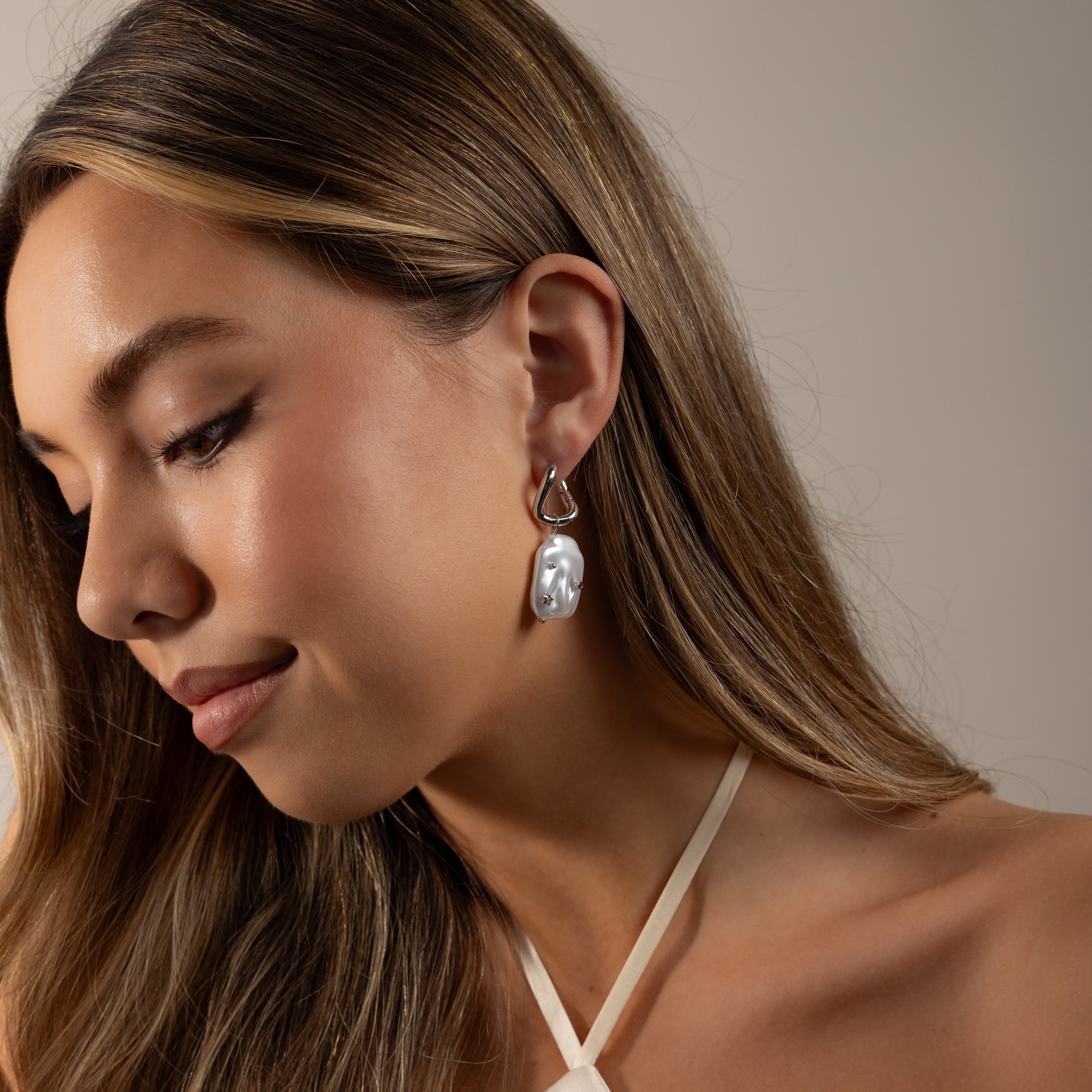 Woman with long brown hair wearing a white halter top and Celestial Baroque Pearl Charm Hoops, looking downward.