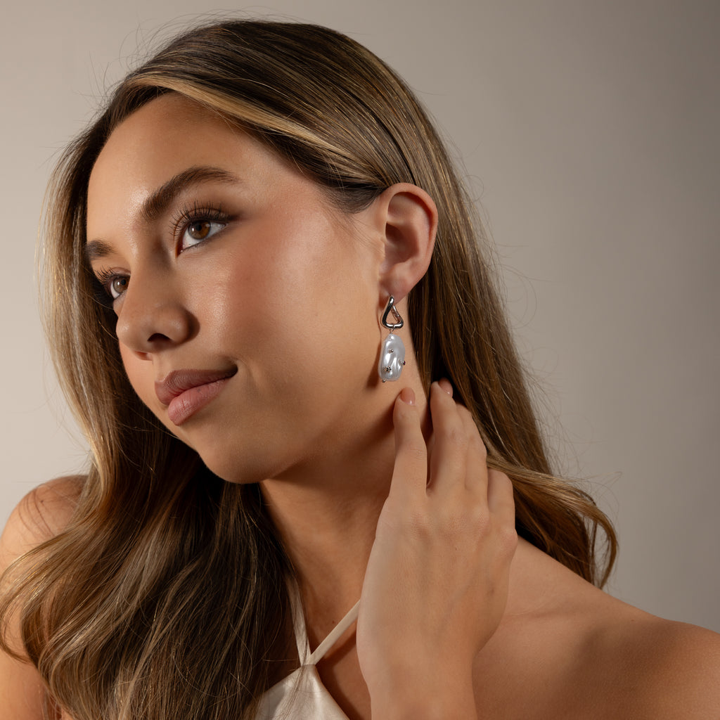 Woman with long hair in a white top holds the Celestial Baroque Pearl Charm Hoops in White Gold, gazing slightly upward.