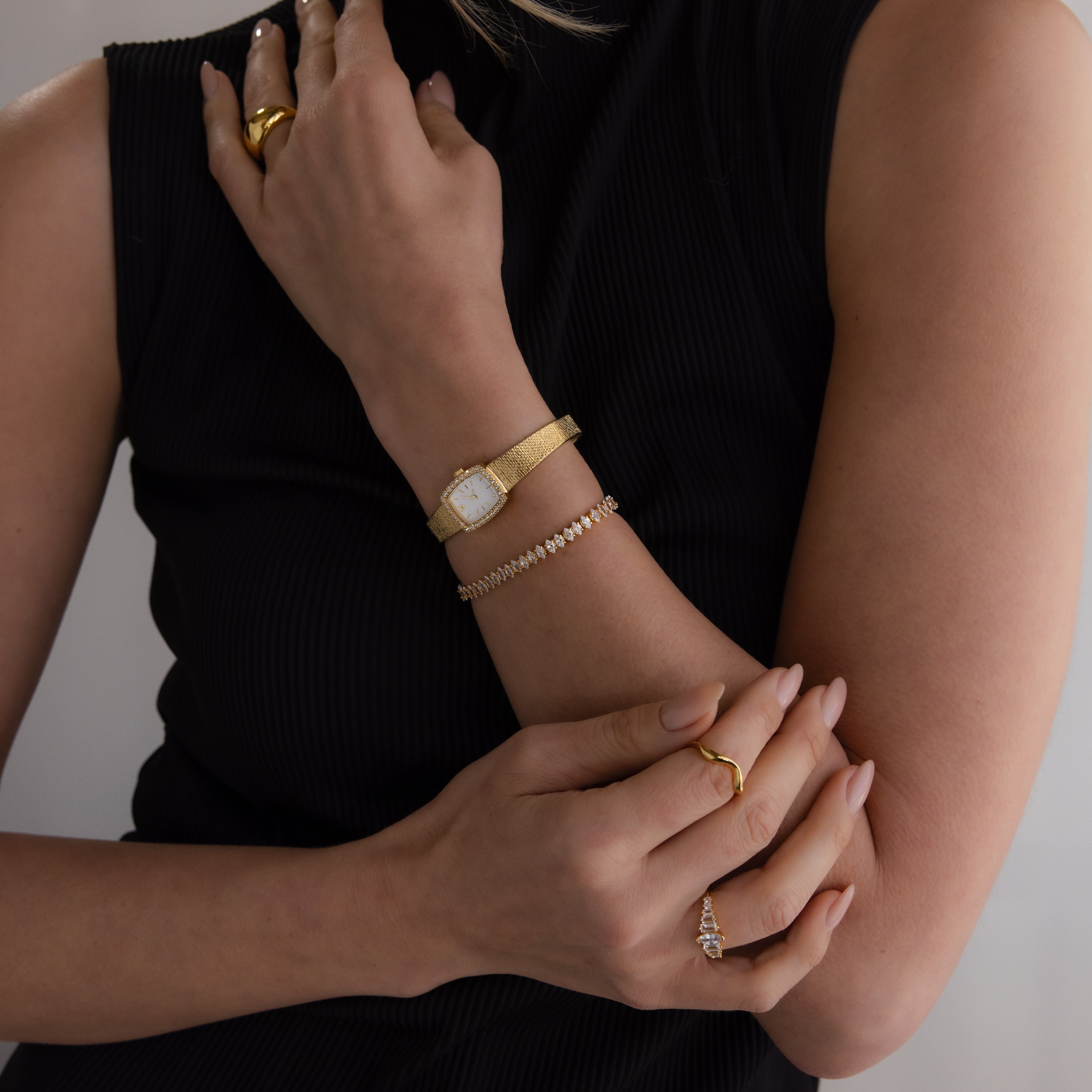 Woman in a sleeveless black top wears gold rings, a delicate bracelet, and the Petite Mesh Timepiece diamond bezel watch.