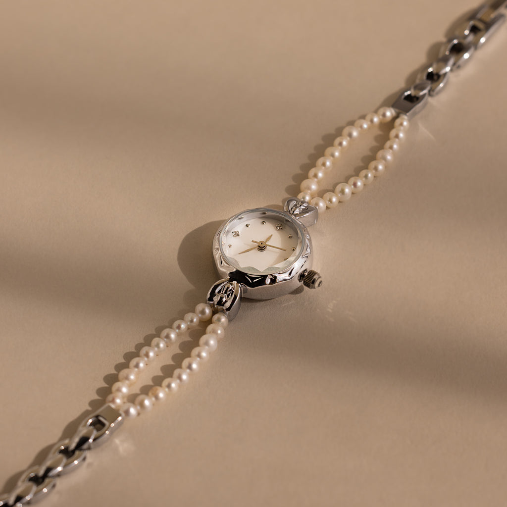 The Beaded Pearl Timepiece in Sterling Silver features a round face and a pearl-beaded strap, offering classic elegance and timeless charm on a beige background.