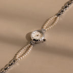 The Beaded Pearl Timepiece in Sterling Silver features a round face and a pearl-beaded strap, offering classic elegance and timeless charm on a beige background.