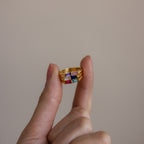 A hand displays three stacked Toi et Moi Tapered Birthstone Rings with vibrant gemstones, including a baguette-cut birthstone, set against a plain beige background.