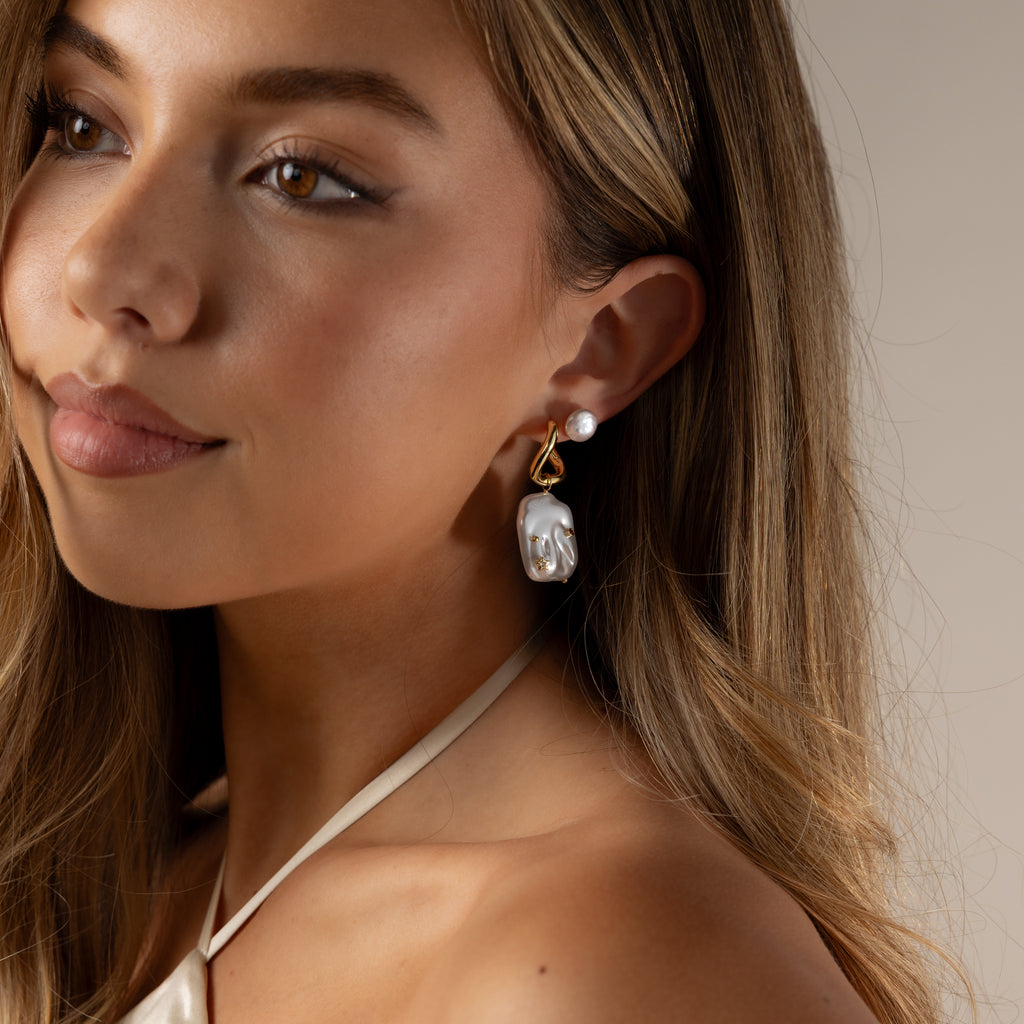 A young woman with long hair wears the Celestial Baroque Pearl Charm Hoops and a cream halter top, gazing slightly to the side.