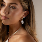 A young woman with long hair wears the Celestial Baroque Pearl Charm Hoops and a cream halter top, gazing slightly to the side.