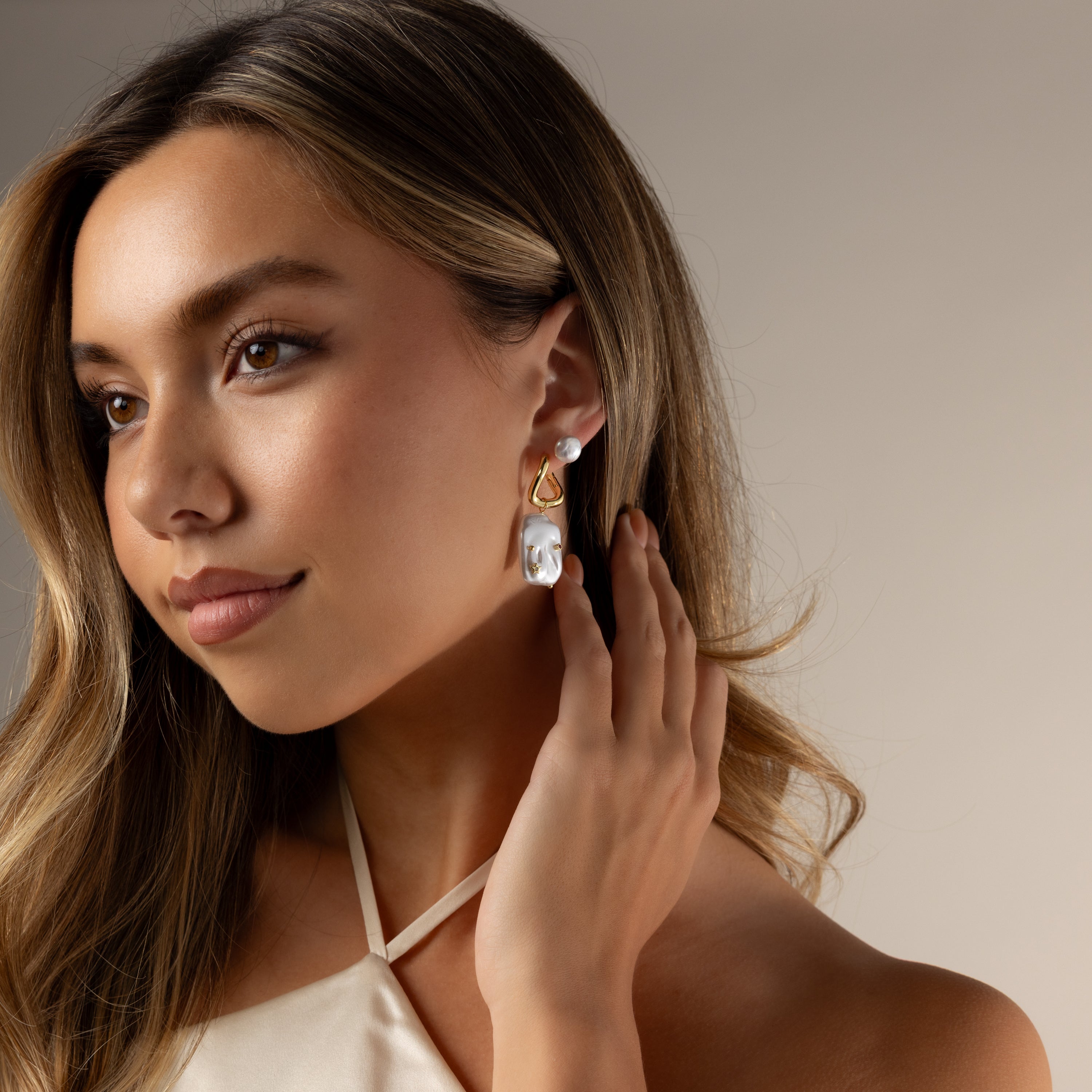 Woman with long hair touches her ear while wearing Celestial Baroque Pearl Charm Hoops and a cream halter top.