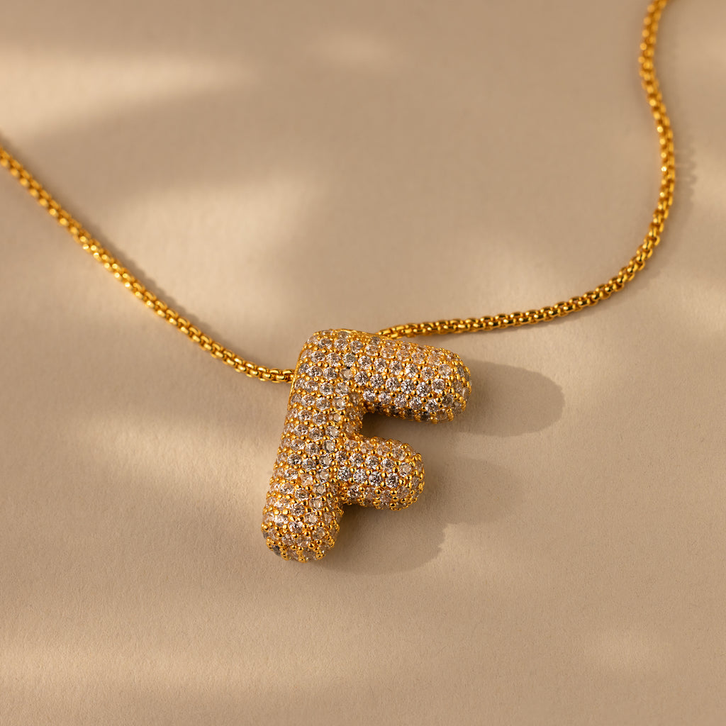 The Pave Puff Letter Necklace in 18K Gold features a pendant shaped like the letter F with pave diamonds, shown on a beige background.