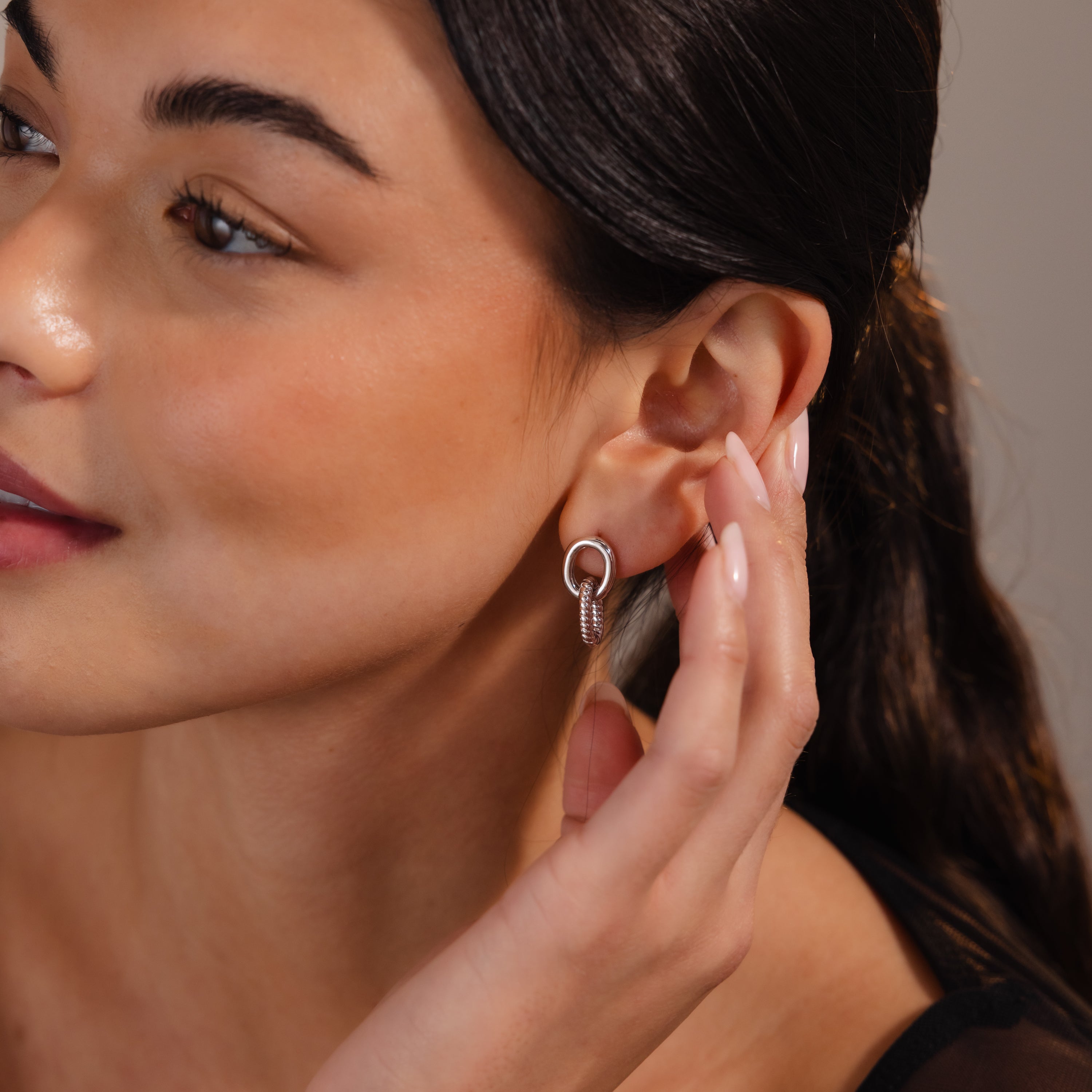 A woman with long dark hair touches her earring, showcasing the Twisted Link Earrings that add a touch of modern jewelry elegance.