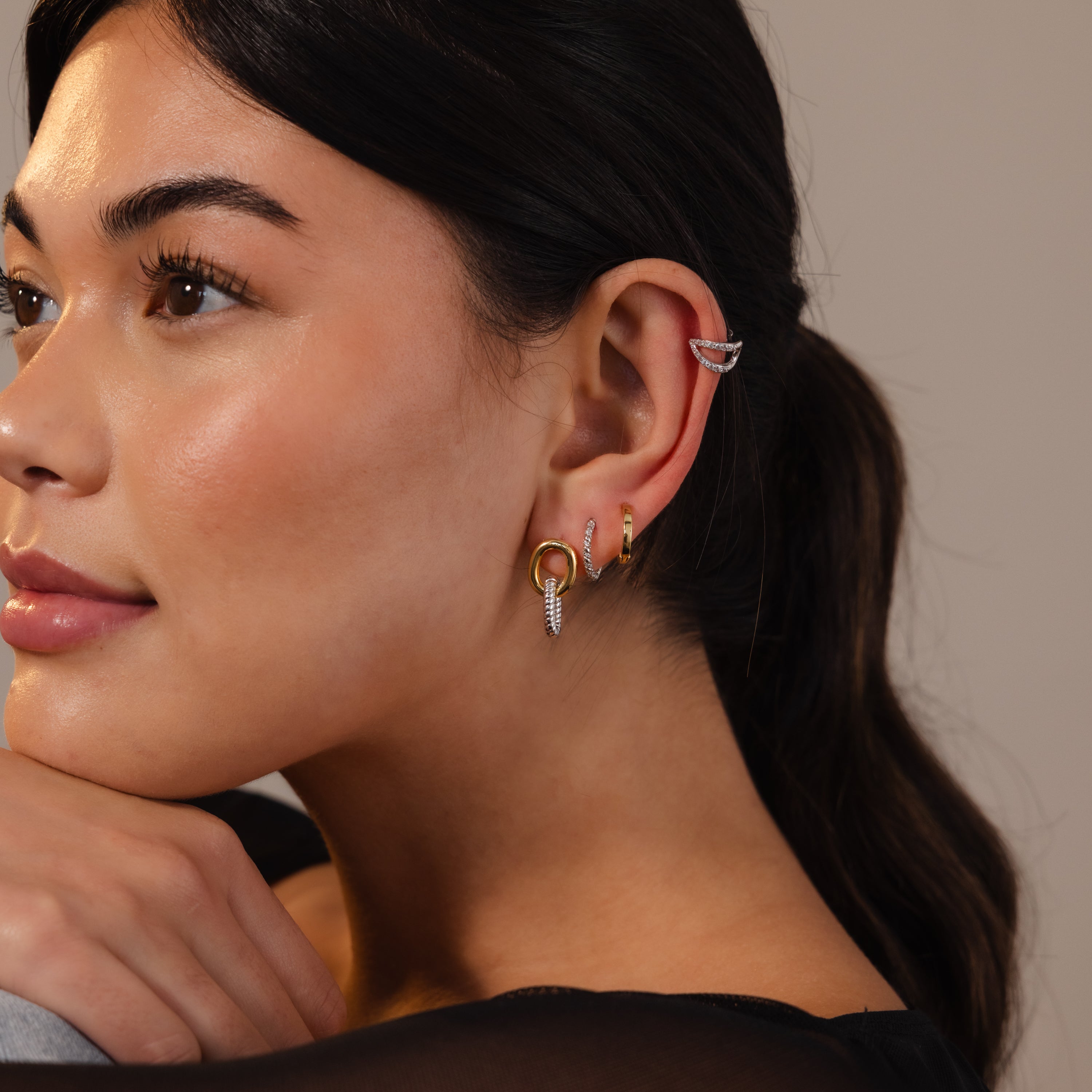 Woman with long dark hair wears multiple modern gold and silver earrings, including the Twisted Link Earrings in Mixed Metal, as she gazes thoughtfully to the side.
