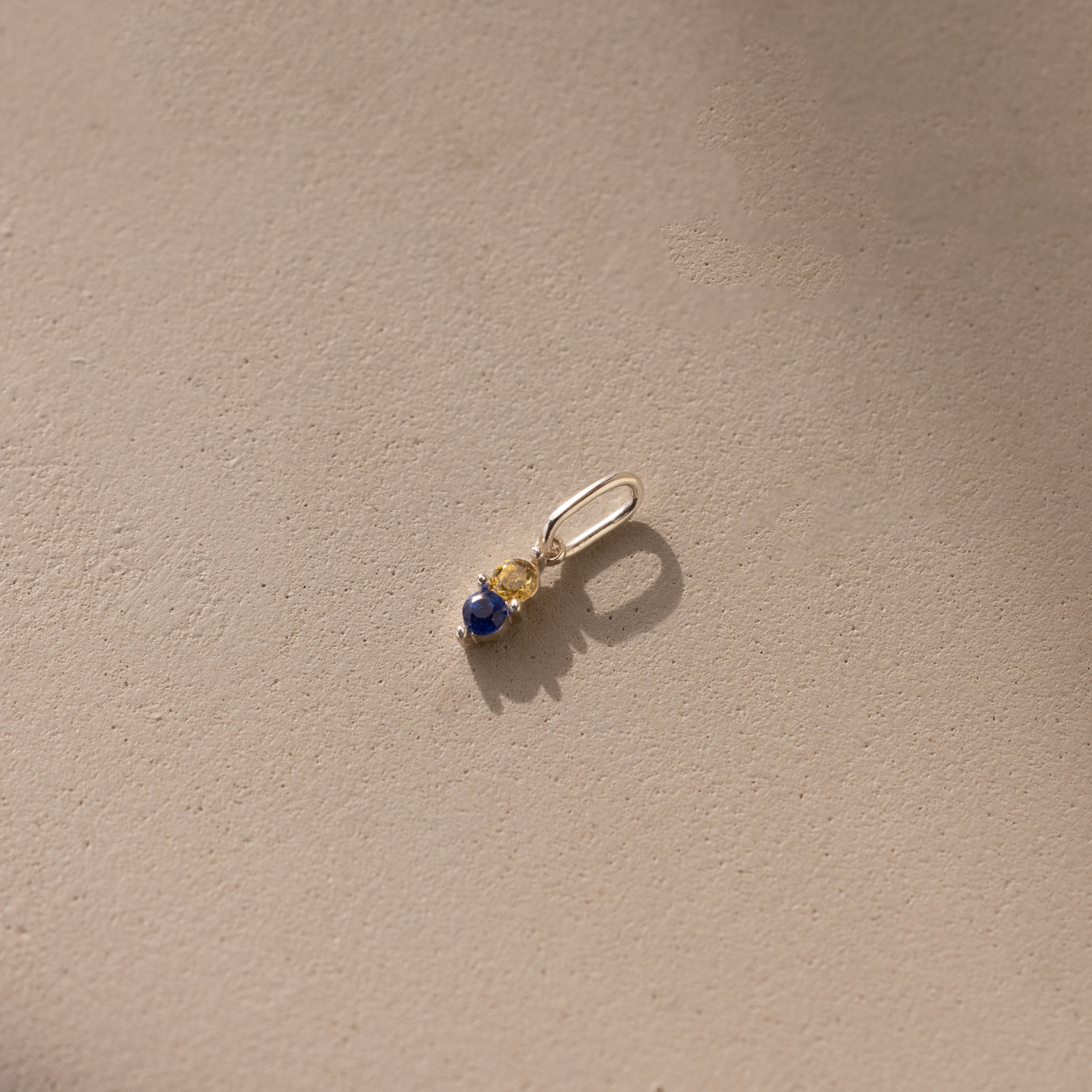Silver Dainty Duo Birthstone Charm with topaz and sapphire. The design features two birthstones stacked vertically in prong setting with a large jump ring attached to the top. 
