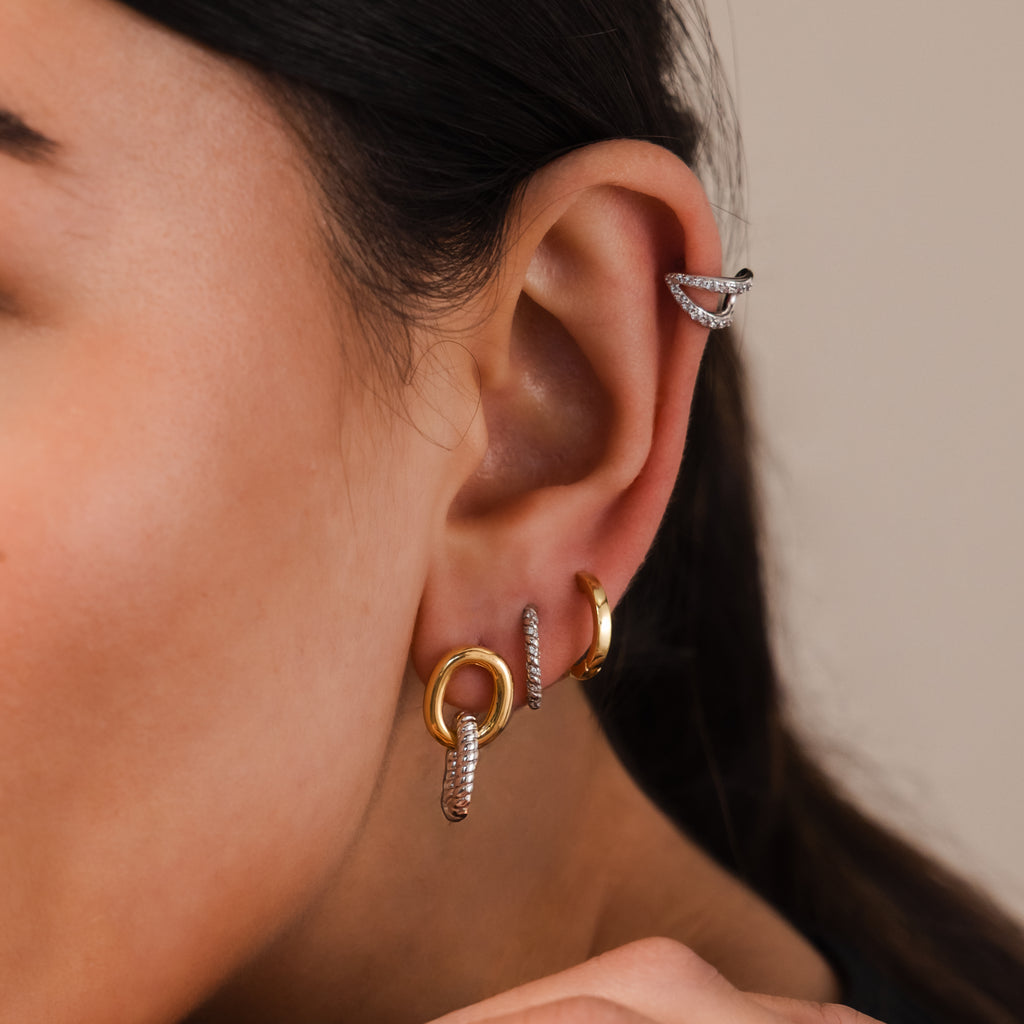Close-up of a person's ear wearing Twisted Link Earrings in Mixed Metal, showcasing modern gold and silver hoops, an ear cuff, and sculptural jewelry accents.