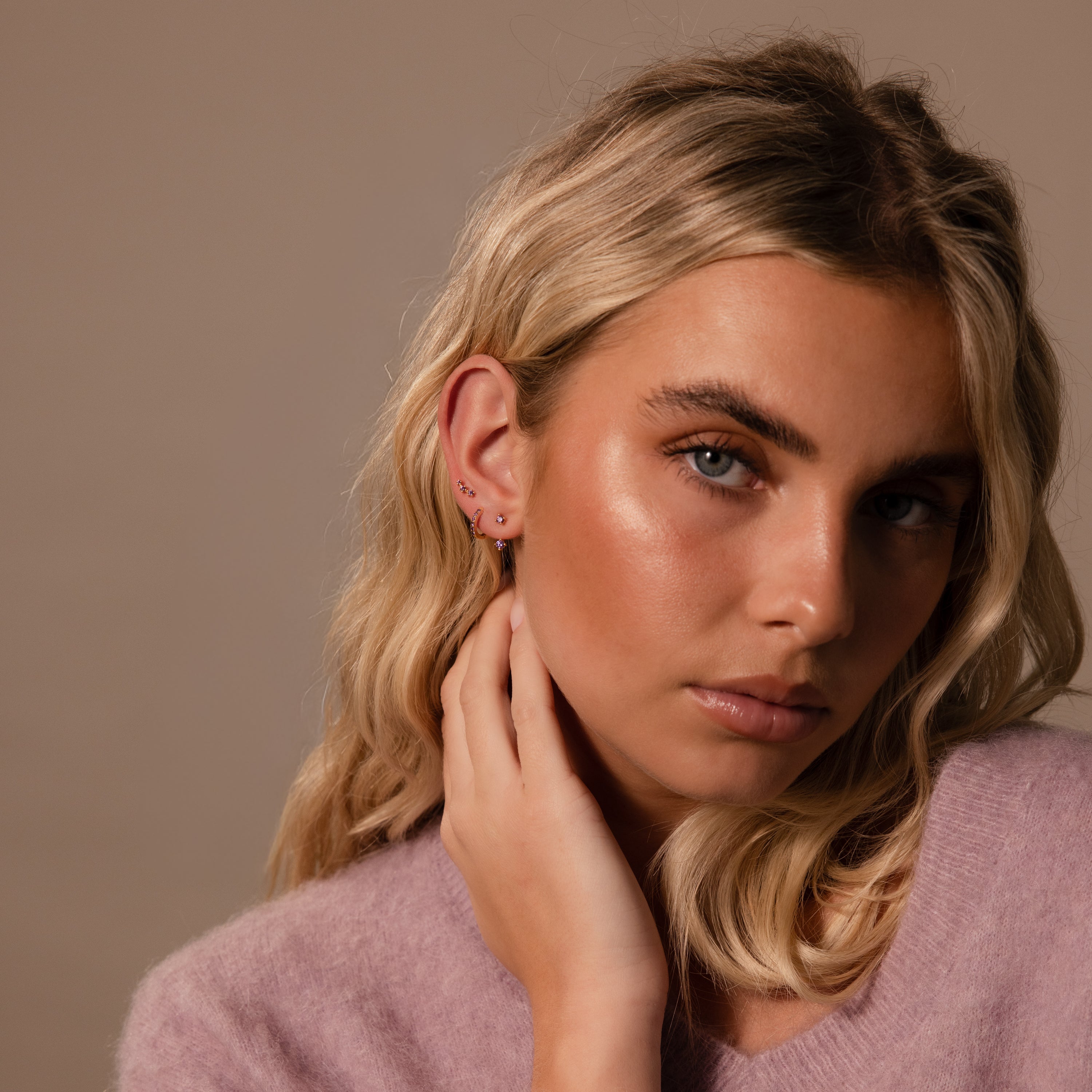 Young woman with wavy blonde hair wearing a lilac sweater, touching her neck and looking at the camera, showcasing the Peridot Earring Trio Set in her ear.
